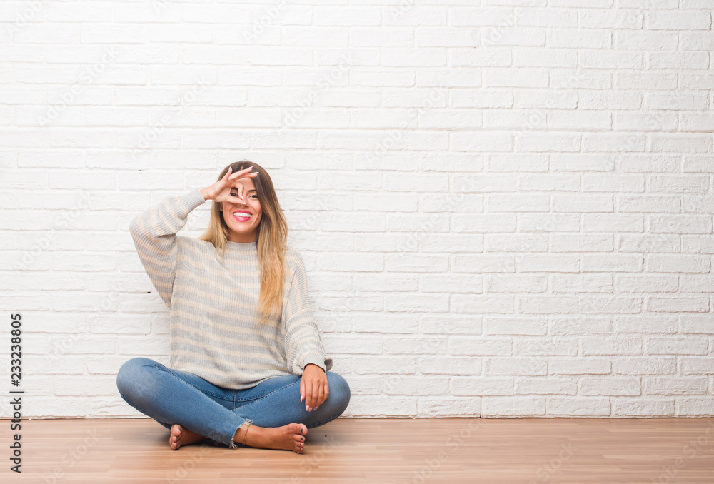 Young adult woman sitting on the floor over white brick wall at home doing ok gesture with hand smiling, eye looking through fingers with happy face. Young adult woman sitting on the floor over white brick wall at home doing ok gesture with hand smiling, eye looking through fingers with happy face.