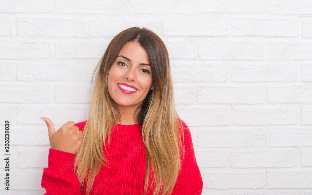 Young adult woman over white brick wall wearing winter outfit at home smiling with happy face looking and pointing to the side with thumb up. Young adult woman over white brick wall wearing winter outfit at home smiling with happy face looking and pointing to the side with thumb up.