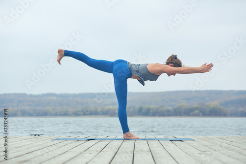 Beautiful young woman practices yoga asana Virabhadrasana 3 - warrior pose 3 on the wooden deck near the lake photo