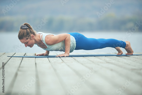 Beautiful young woman practices yoga asana Chaturanga Dandasana - plank pose on the wooden deck near the lake photo