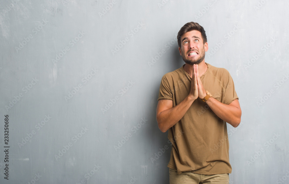 Handsome young man over grey grunge wall begging and praying with hands together with hope expression on face very emotional and worried. Asking for forgiveness. Religion concept.