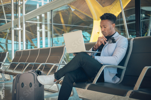 Young businessman is seated in the airport working with a laptop and carrying a suitcase waiting for his flight