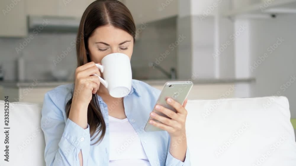 Brunette woman with brown eyes sitting in living room using smartphone. Young female browsing on mobile phone chatting online with friends. Housewife drink coffee from white cup. Kitchen on background Brunette woman with brown eyes sitting in living room using smartphone. Young female browsing on mobile phone chatting online with friends. Housewife drink coffee from white cup. Kitchen on background