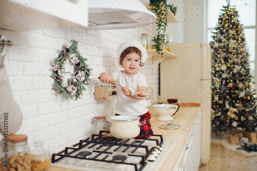Cute little girl in santa hat, preparing cookies in the kitchen at home. Sits on the kitchen table and helps mom prepare a festive Christmas dinner