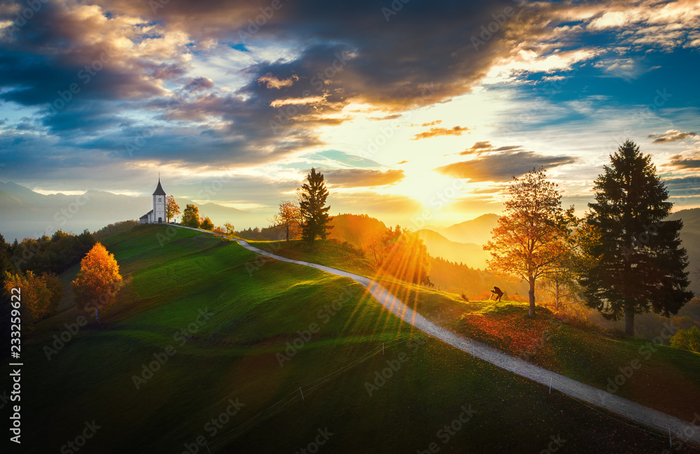 Fototapeta Jamnik, Słowenia - Widok z lotu ptaka na kościół St. Primoz w Słowenii w pobliżu Jamnika z pięknymi chmurami i Alpami Julijskimi w tle.