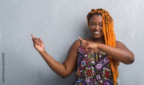 Young african american woman over grey grunge wall wearing orange braids smiling and looking at the camera pointing with two hands and fingers to the side. photo