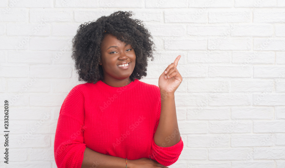 Young african american plus size woman over white brick wall with a big smile on face, pointing with hand and finger to the side looking at the camera. Young african american plus size woman over white brick wall with a big smile on face, pointing with hand and finger to the side looking at the camera.