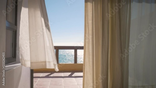 Balcony of a tropical luxury hotel by the ocean in 4k. Inside looking out at white curtains blowing in a warm summer wind with the ocean stretching out in the background. Wide shot depicts wanderlust.