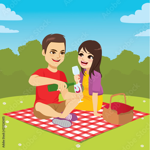 Cute young couple on picnic day enjoying wine sitting on patterned blanket at park with basket