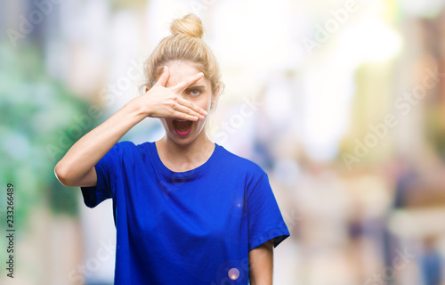 Young beautiful blonde and blue eyes woman wearing blue t-shirt over isolated background peeking in shock covering face and eyes with hand, looking through fingers with embarrassed expression.