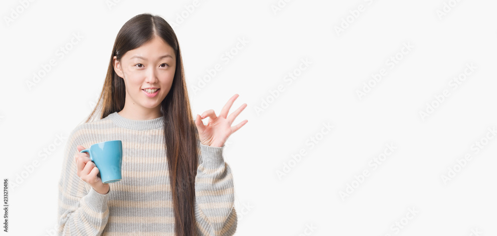 Young Chinese woman over isolated background driking cup of coffee doing ok sign with fingers, excellent symbol Young Chinese woman over isolated background driking cup of coffee doing ok sign with fingers, excellent symbol