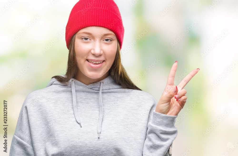 Young caucasian beautiful woman wearing wool cap over isolated background smiling with happy face winking at the camera doing victory sign. Number two. Young caucasian beautiful woman wearing wool cap over isolated background smiling with happy face winking at the camera doing victory sign. Number two.