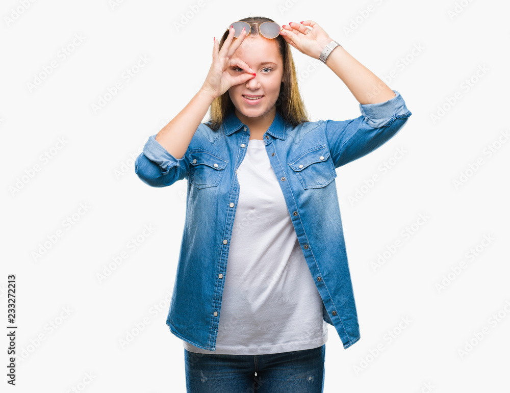 Young beautiful caucasian woman wearing sunglasses over isolated background doing ok gesture with hand smiling, eye looking through fingers with happy face. Young beautiful caucasian woman wearing sunglasses over isolated background doing ok gesture with hand smiling, eye looking through fingers with happy face.