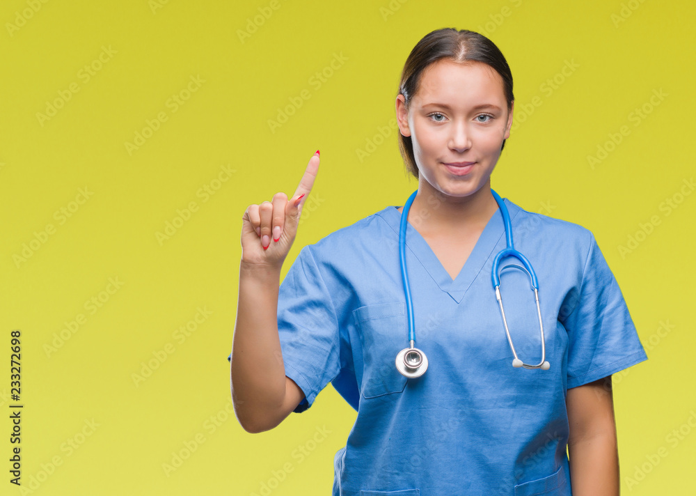Young caucasian doctor woman wearing medical uniform over isolated background showing and pointing up with finger number one while smiling confident and happy. Young caucasian doctor woman wearing medical uniform over isolated background showing and pointing up with finger number one while smiling confident and happy.
