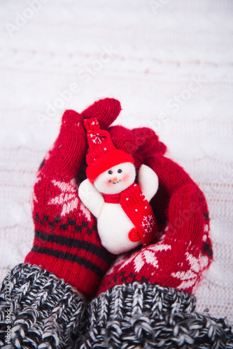 Hands in red winter gloves holding toy cute snowman on white knitted background.