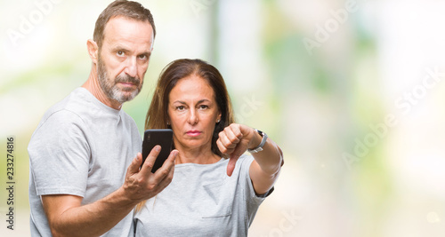 Middle age hispanic couple texting message on smartphone ver isolated background with angry face, negative sign showing dislike with thumbs down, rejection concept