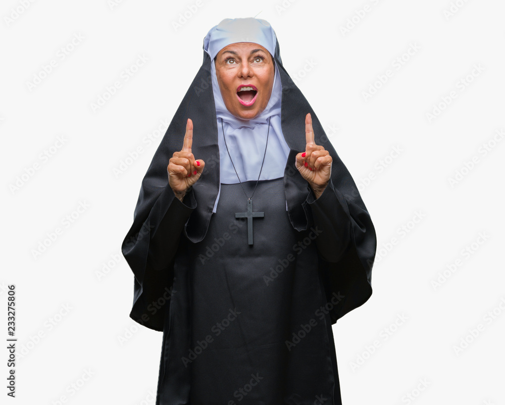 Middle age senior christian catholic nun woman over isolated background amazed and surprised looking up and pointing with fingers and raised arms. Middle age senior christian catholic nun woman over isolated background amazed and surprised looking up and pointing with fingers and raised arms.