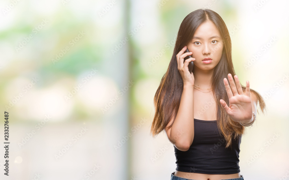 Young asian woman speaking on the phone over isolated background with open hand doing stop sign with serious and confident expression, defense gesture Young asian woman speaking on the phone over isolated background with open hand doing stop sign with serious and confident expression, defense gesture