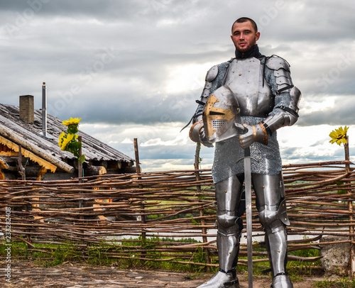 Knight in armor on the background of a wicker fence, a wooden house and the expanses of the motherland. Knightly armor and weapon. Semi - antique photo.