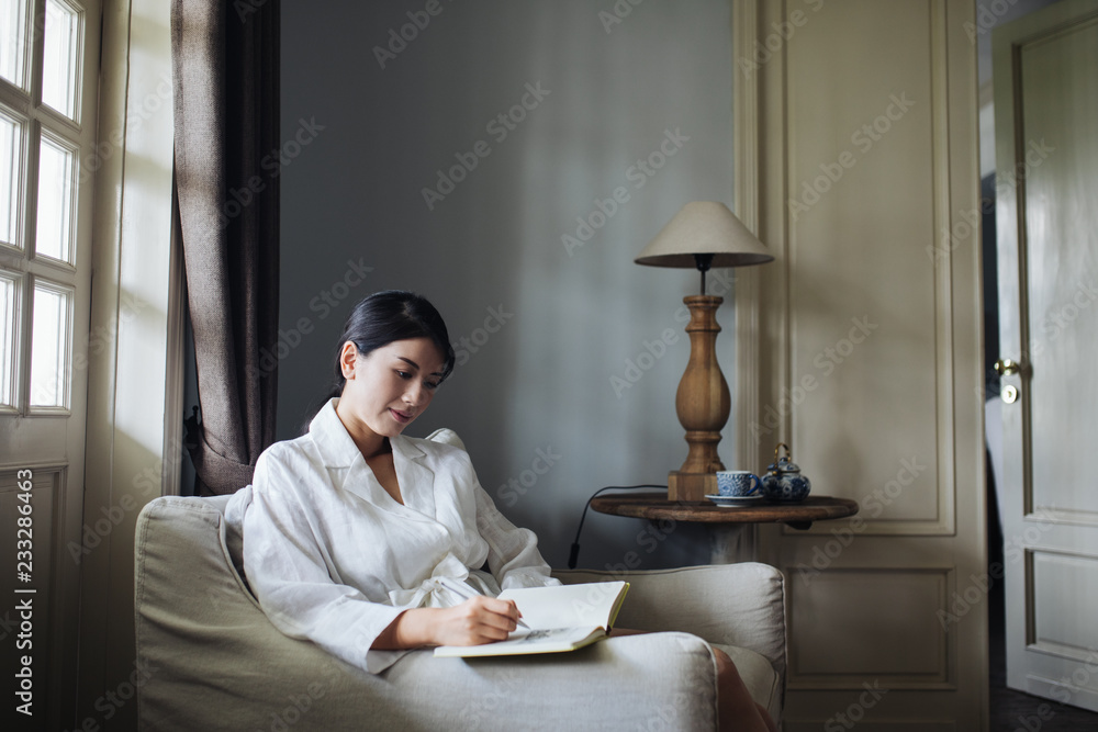 © LUMINA IMAGES - Young woman writing in notebook while sitting on armchair at home © LUMINA IMAGES - Young woman writing in notebook while sitting on armchair at home