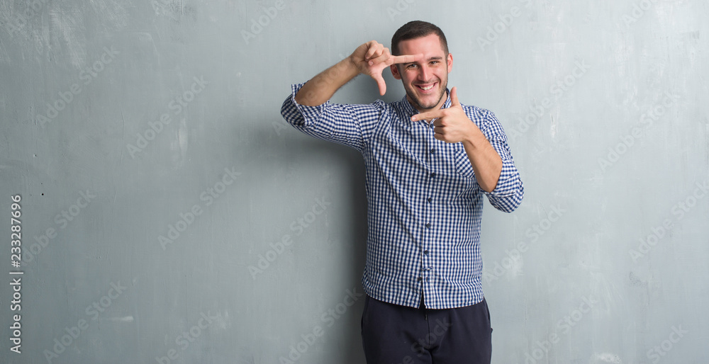 Young caucasian man over grey grunge wall smiling making frame with hands and fingers with happy face. Creativity and photography concept.