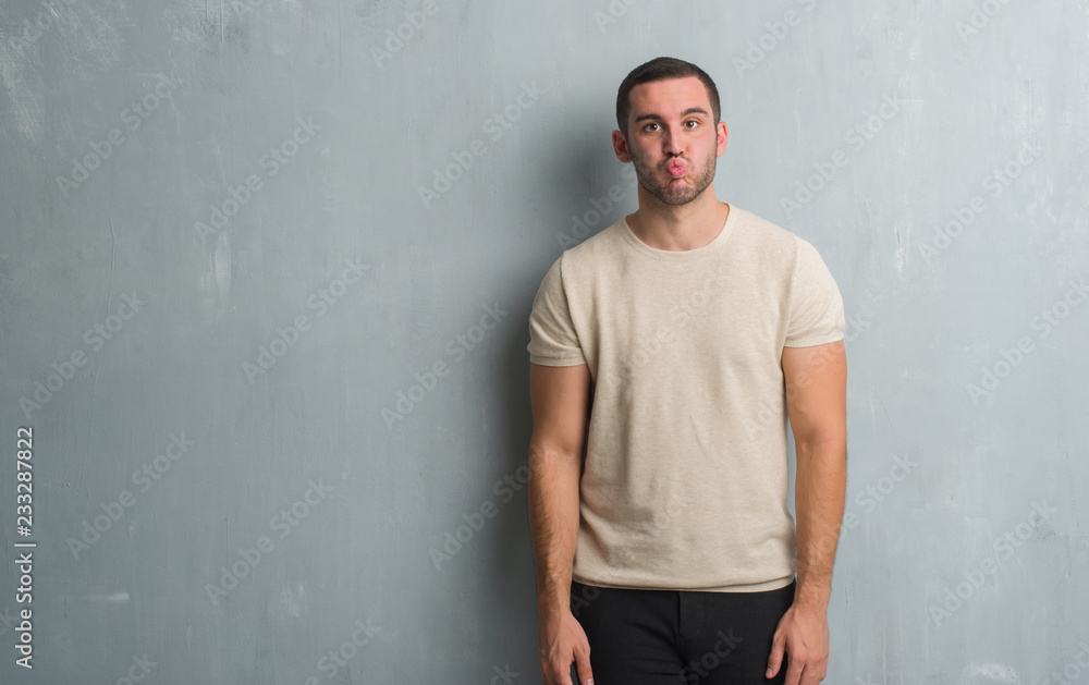 Young caucasian man over grey grunge wall puffing cheeks with funny face. Mouth inflated with air, crazy expression. Young caucasian man over grey grunge wall puffing cheeks with funny face. Mouth inflated with air, crazy expression.