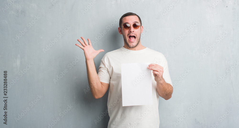 Young caucasian man over grey grunge wall holding blank paper very happy and excited, winner expression celebrating victory screaming with big smile and raised hands Young caucasian man over grey grunge wall holding blank paper very happy and excited, winner expression celebrating victory screaming with big smile and raised hands