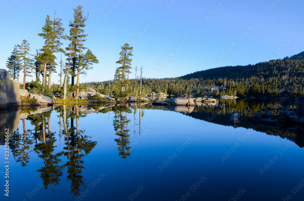 Pine trees, rocks, and perfect blue sky reflected in the serene, calm, and peaceful waters of an alpine lake at sunrise - Middle Velma Lake in the Desolation Wilderness near Lake Tahoe, California