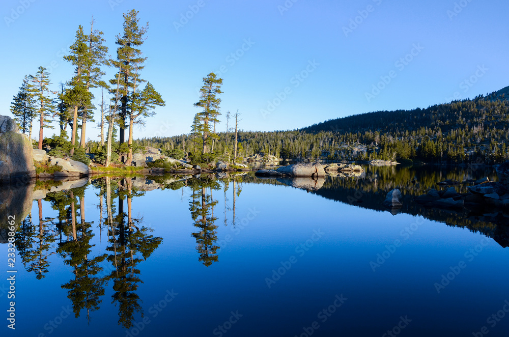 Obraz premium Pine trees, rocks, and perfect blue sky reflected in the serene, calm, and peaceful waters of an alpine lake at sunrise - Middle Velma Lake in the Desolation Wilderness near Lake Tahoe, California
