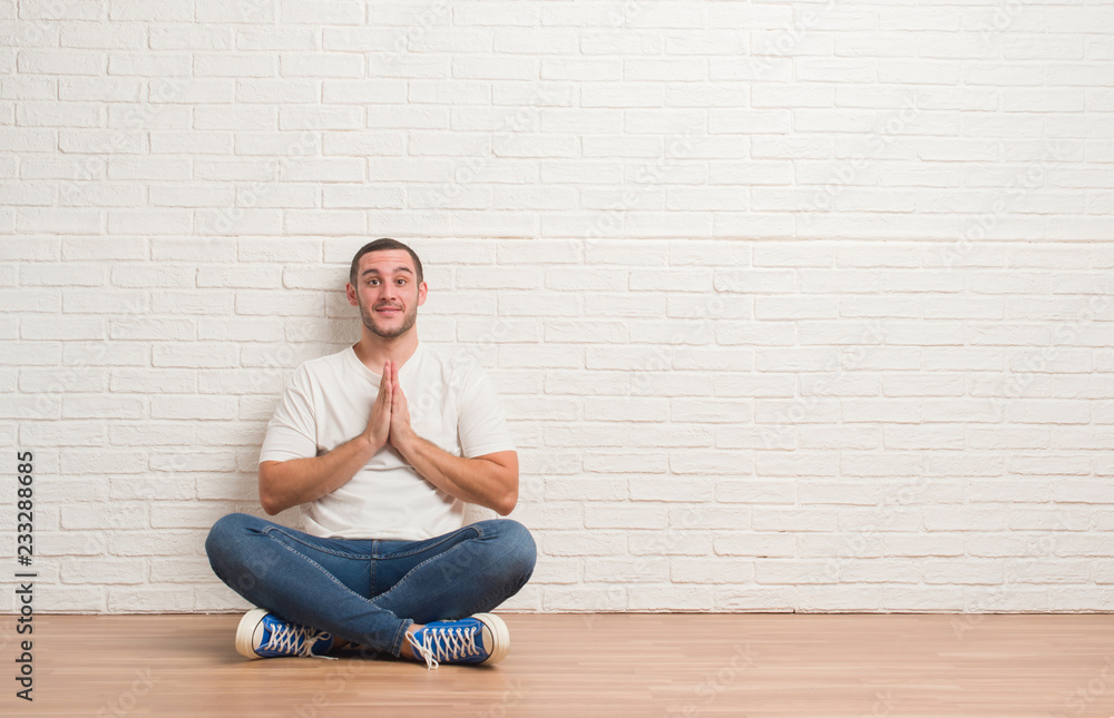 Young caucasian man sitting on the floor over white brick wall praying with hands together asking for forgiveness smiling confident. Young caucasian man sitting on the floor over white brick wall praying with hands together asking for forgiveness smiling confident.