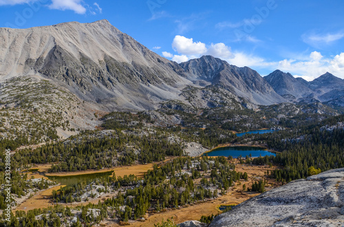 View of granite mountain peaks, alpine valley, meadows, and lakes from a high overlook under a bright blue sky with puffy white clouds - Sierra Nevada Mountains and Little Lakes Valley in California
