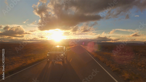 AERIAL: Black SUV car driving along the empty countryside road at golden sunset
