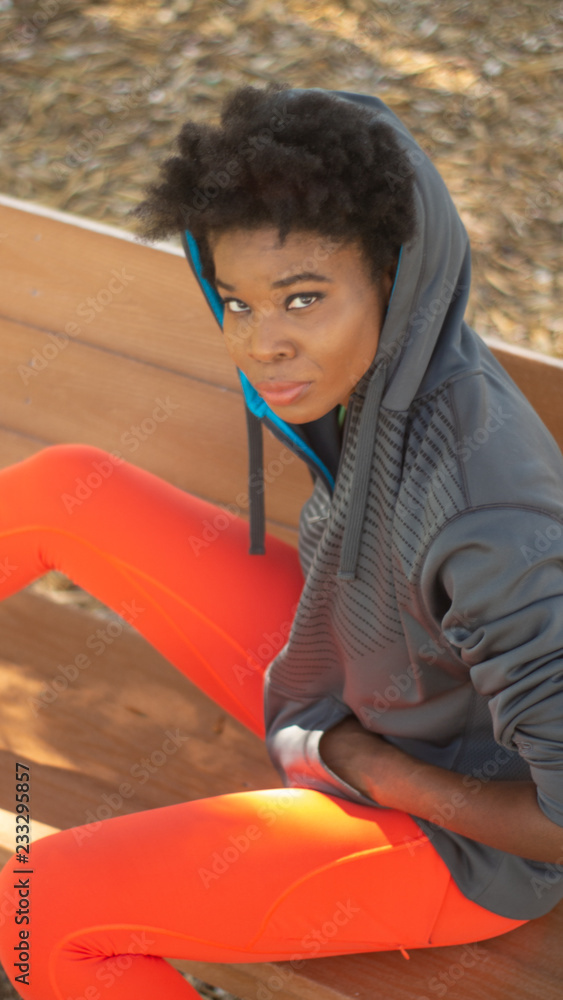 Strong african american woman in orange running tights and gray and blue sweater resting on a bench during a cool fall day