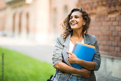 Pretty cheerful latin american student smiling at camera carrying notebook on campus at college photo