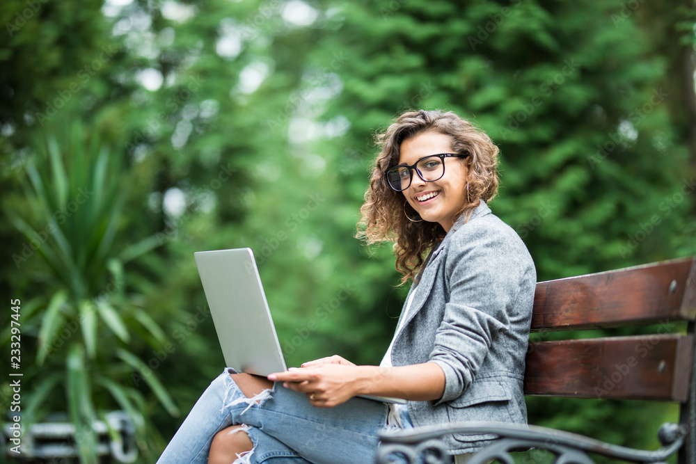Young female student in eyesglasses sitting on a bench in park, she is working on laptop. Young female student in eyesglasses sitting on a bench in park, she is working on laptop.
