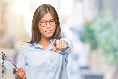 Young asian business woman over isolated background holding clipboard pointing with finger to the camera and to you, hand sign, positive and confident gesture from the front