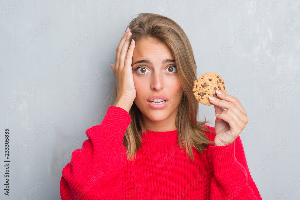 Beautiful young woman over grunge grey wall eating chocolate chip cooky stressed with hand on head, shocked with shame and surprise face, angry and frustrated. Fear and upset for mistake. Beautiful young woman over grunge grey wall eating chocolate chip cooky stressed with hand on head, shocked with shame and surprise face, angry and frustrated. Fear and upset for mistake.