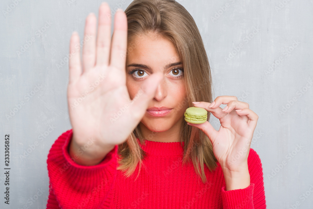 Beautiful young woman over grunge grey wall eating green macaron with open hand doing stop sign with serious and confident expression, defense gesture Beautiful young woman over grunge grey wall eating green macaron with open hand doing stop sign with serious and confident expression, defense gesture