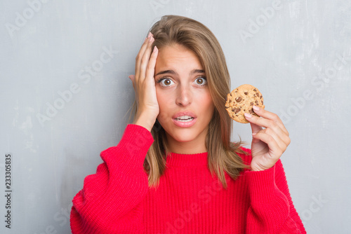 Beautiful young woman over grunge grey wall eating chocolate chip cooky stressed with hand on head  shocked with shame and surprise face  angry and frustrated. Fear and upset for mistake.