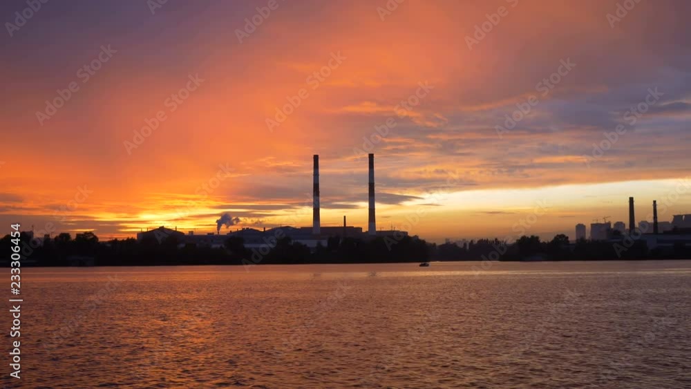 Incredible view of the evening city from the ship on the river in the background of the sunset. Silhouette of large pipes of the plant against the sky. Production by the river at the edge of the city. Incredible view of the evening city from the ship on the river in the background of the sunset. Silhouette of large pipes of the plant against the sky. Production by the river at the edge of the city.