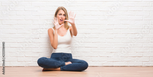 Beautiful young woman sitting on the floor at home afraid and terrified with fear expression stop gesture with hands, shouting in shock. Panic concept.