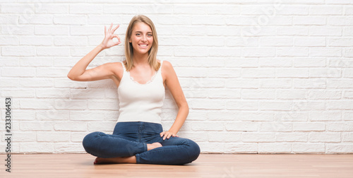 Beautiful young woman sitting on the floor at home smiling positive doing ok sign with hand and fingers. Successful expression.