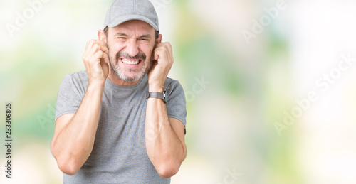 Handsome middle age hoary senior man wearing sport cap over isolated background covering ears with fingers with annoyed expression for the noise of loud music. Deaf concept.