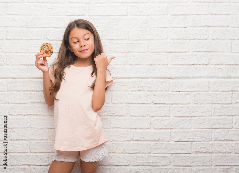 Young hispanic kid over white brick wall eating chocolate chips cooky pointing and showing with thumb up to the side with happy face smiling Young hispanic kid over white brick wall eating chocolate chips cooky pointing and showing with thumb up to the side with happy face smiling