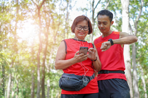 Happy senior asian woman with man or personal trainer checking time from smart watch. Checking heart rate while jogging in the park. Looking on smartwatch her heartbeat while running, Elderly care