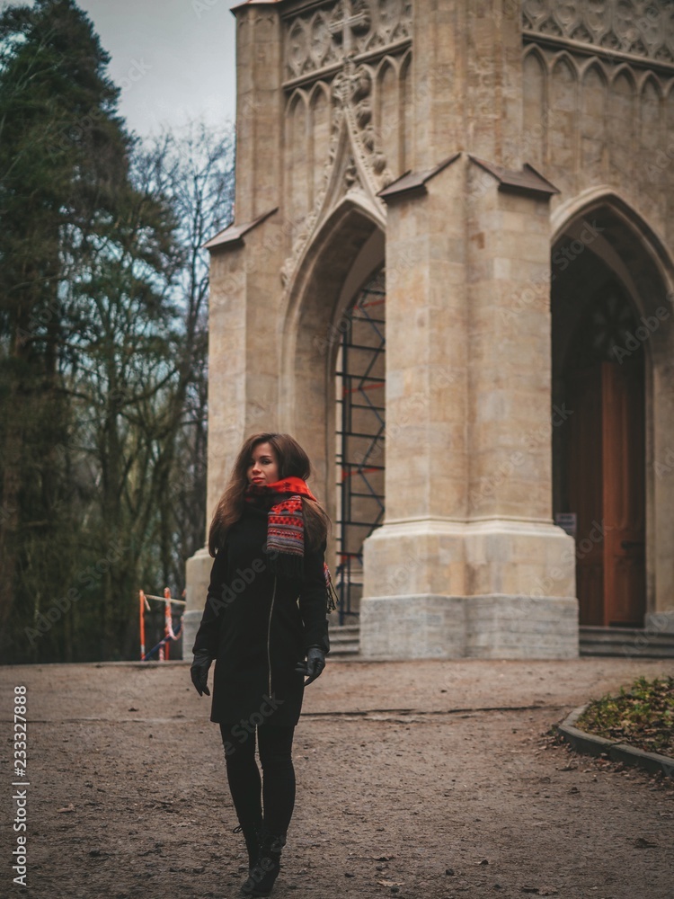 Brunette girl with long hair in black coat and bright red scarf stands near old Gothic Church / Cathedral in the forest
