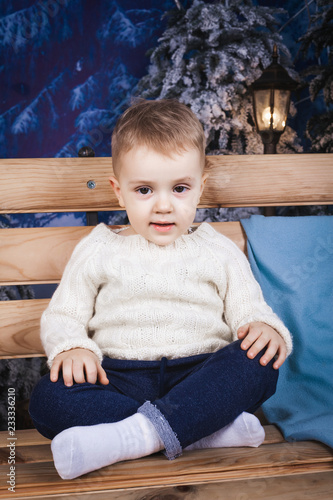 small serious baby boy wearing sweater and jeans sitting on the wooden bench near a blue pillow indoor photo