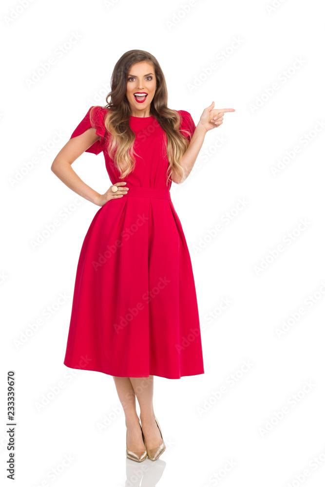 Surprised Fashion Woman In Red Dress And Gold High Heels Is Looking At Camera And Pointing