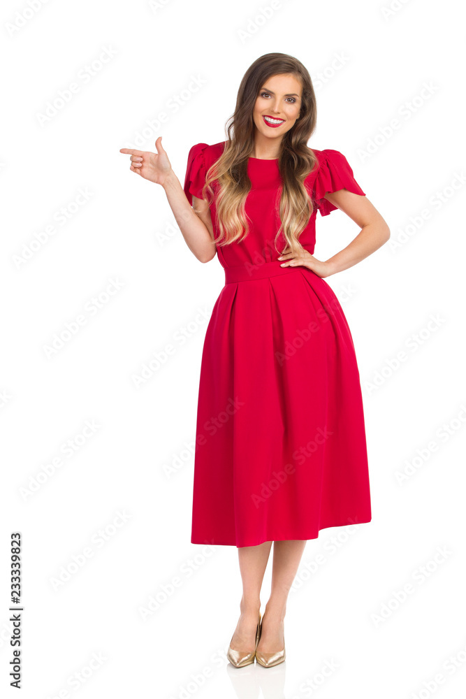 Elegant Woman In Red Dress And Gold High Heels Is Looking At Camera, Smiling And Pointing