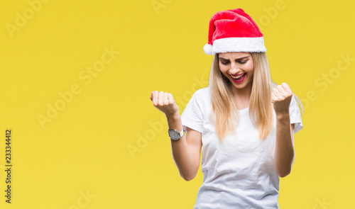 Young beautiful blonde woman christmas hat over isolated background very happy and excited doing winner gesture with arms raised, smiling and screaming for success. Celebration concept. © Krakenimages.com